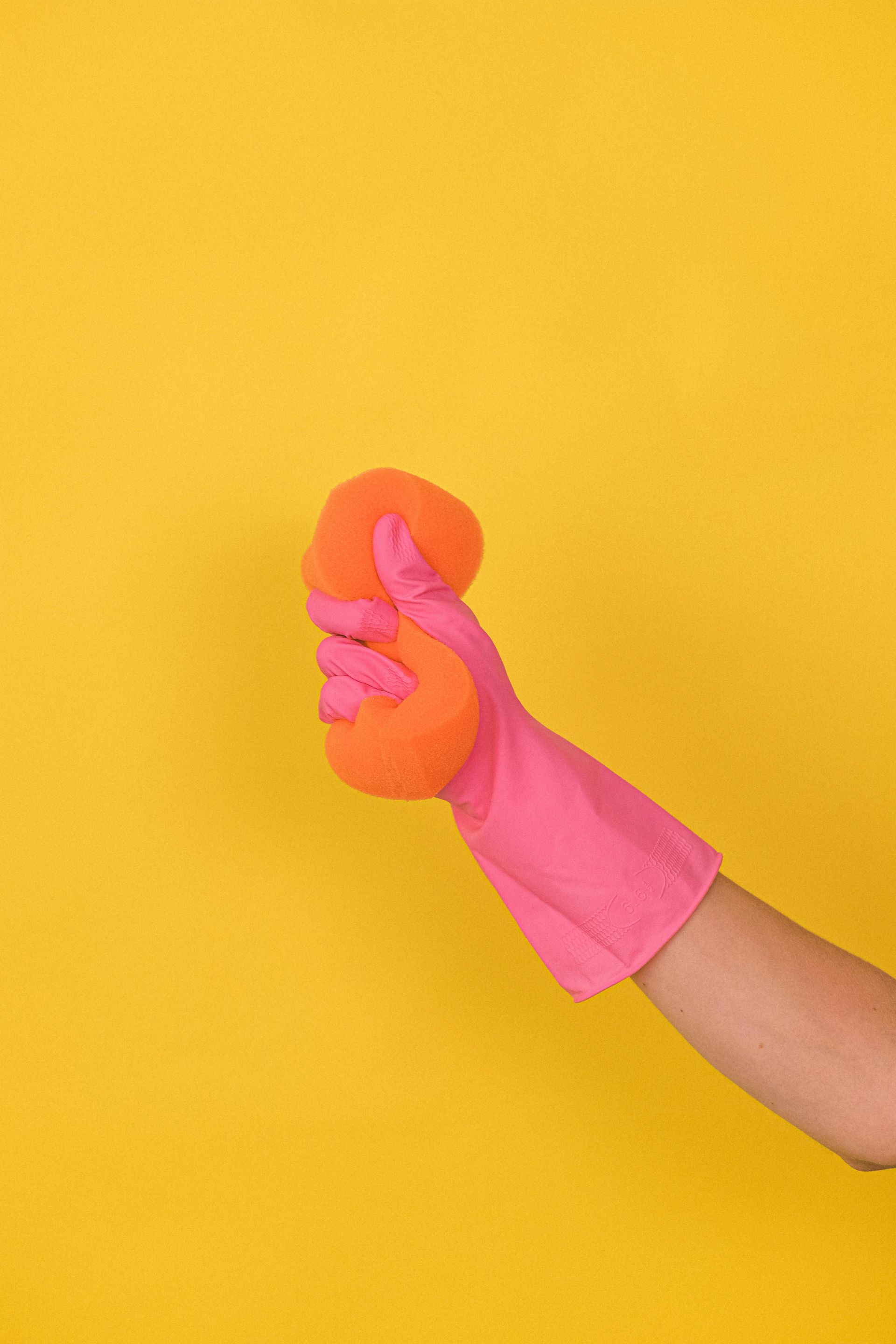 Pink-gloved hand squeezing an orange sponge against a yellow background.