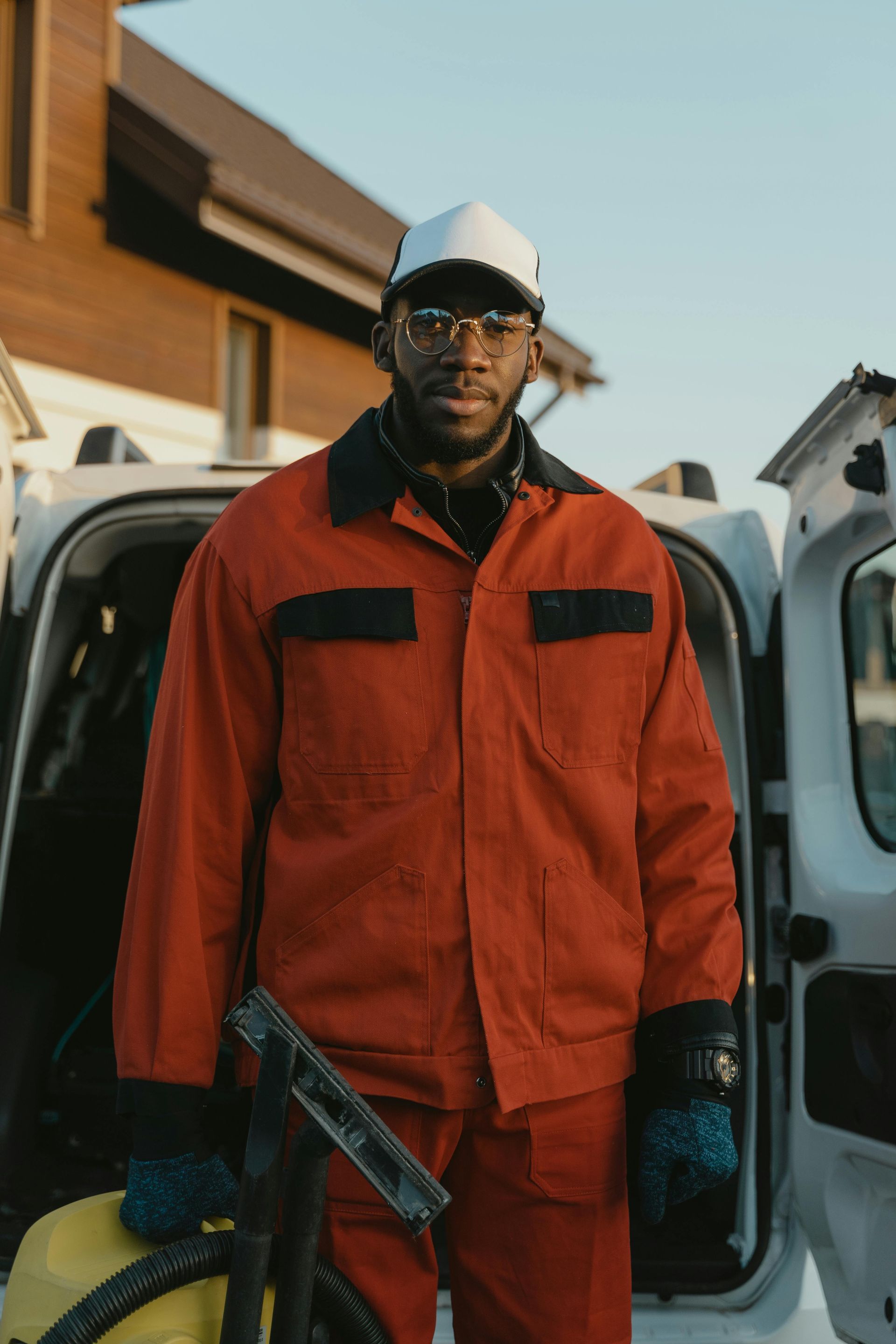 Man in red work suit and white cap, holding cleaning equipment in front of a white van.