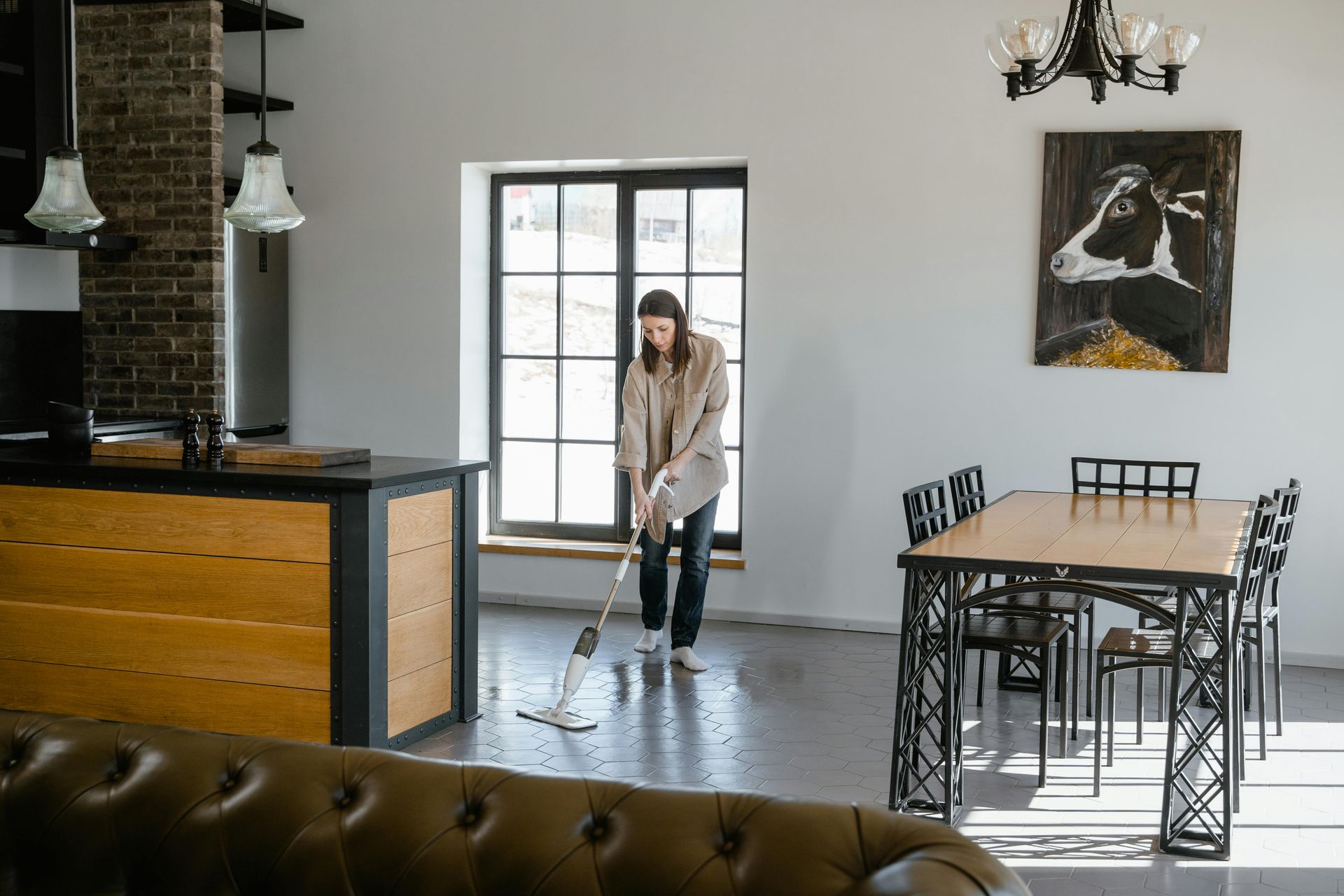 Woman mopping a light-colored floor in a modern kitchen/dining area. Includes a table, chairs, and a cow painting.