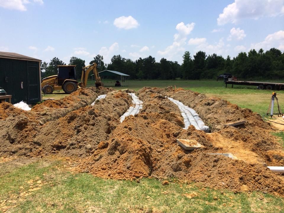 Construction site with trenches, a backhoe, and dirt piles on a grassy field.
