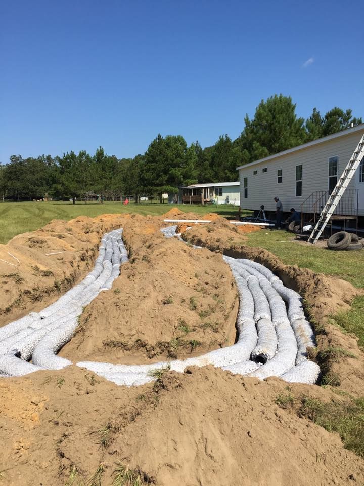 Trenches dug in yard with white septic system components, near a mobile home and trees.
