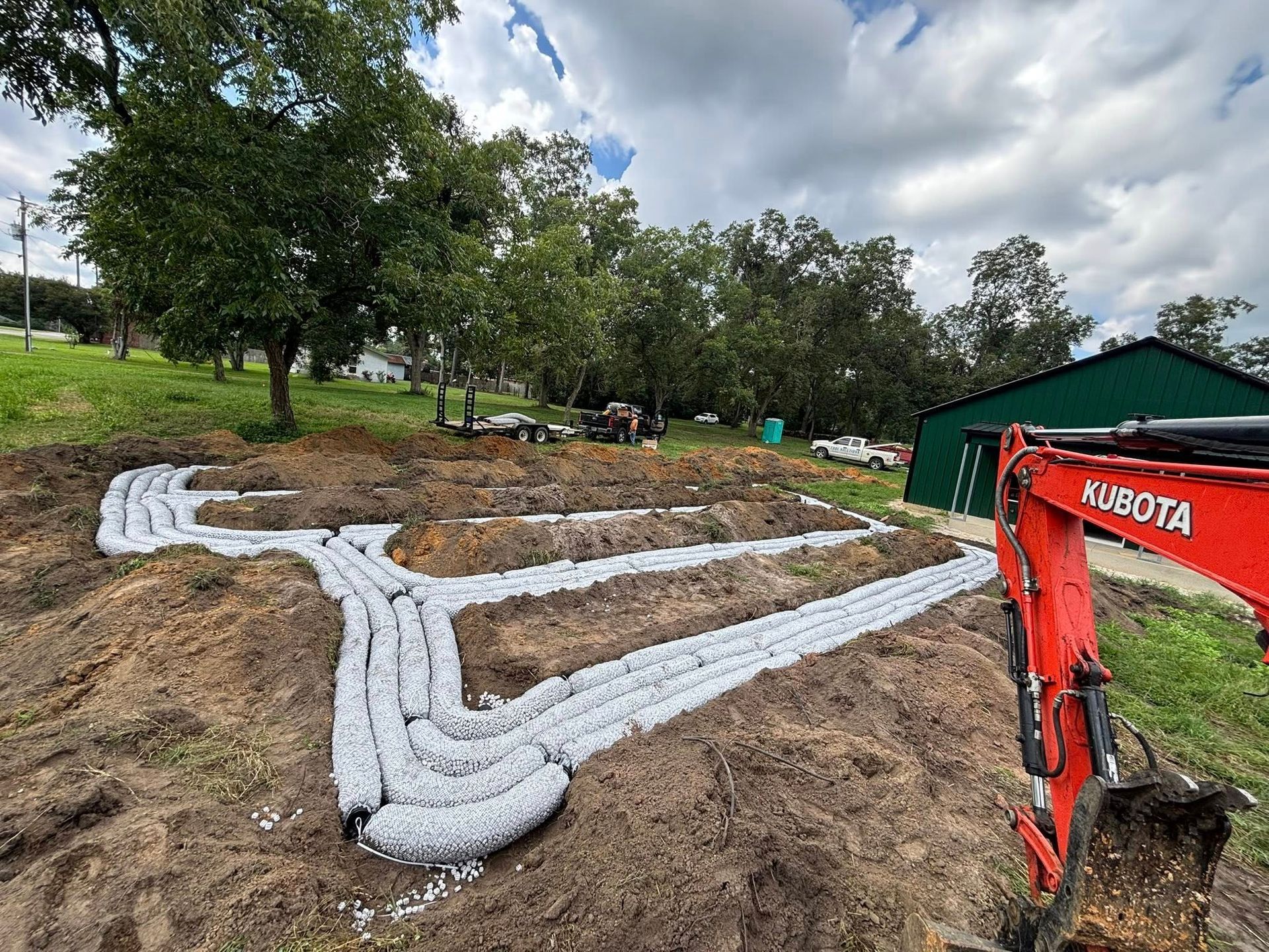 An excavator is creating a drainage system with white gravel in a field with trees and a building.