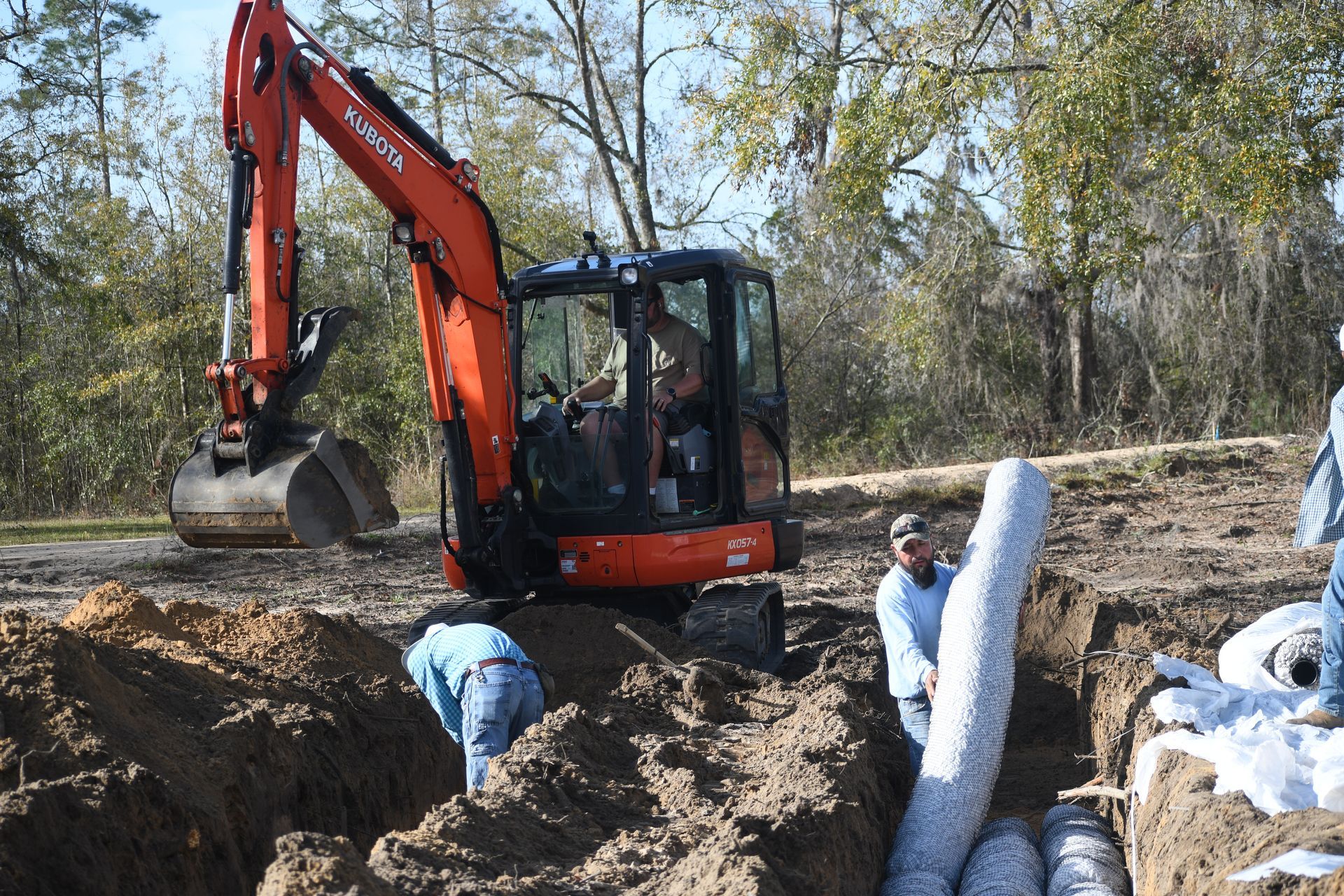 Orange excavator and workers installing large drainage pipes in a muddy trench outdoors