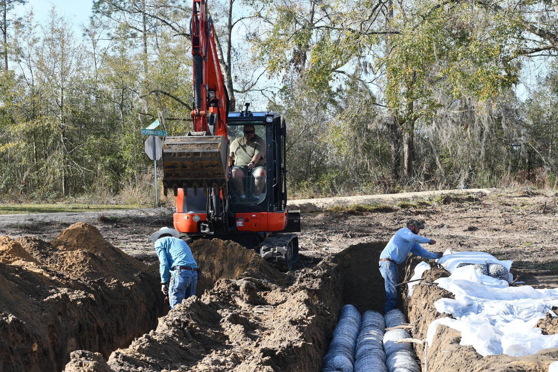 Excavator digging a trench beside stacked white pipes, with two workers in a wooded outdoor worksite.