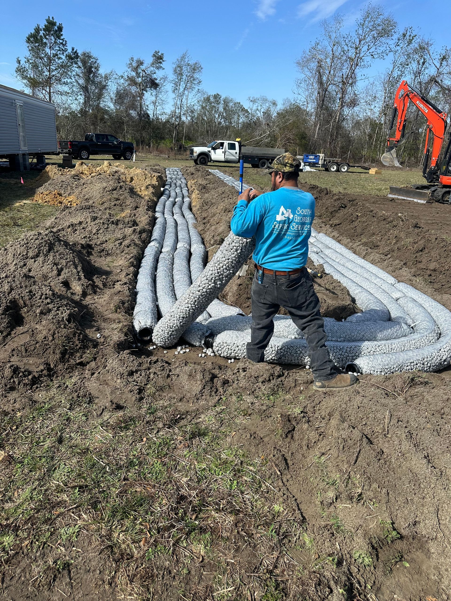 Worker arranging white geotextile fabric in a dirt trench at a construction site with machinery nearby