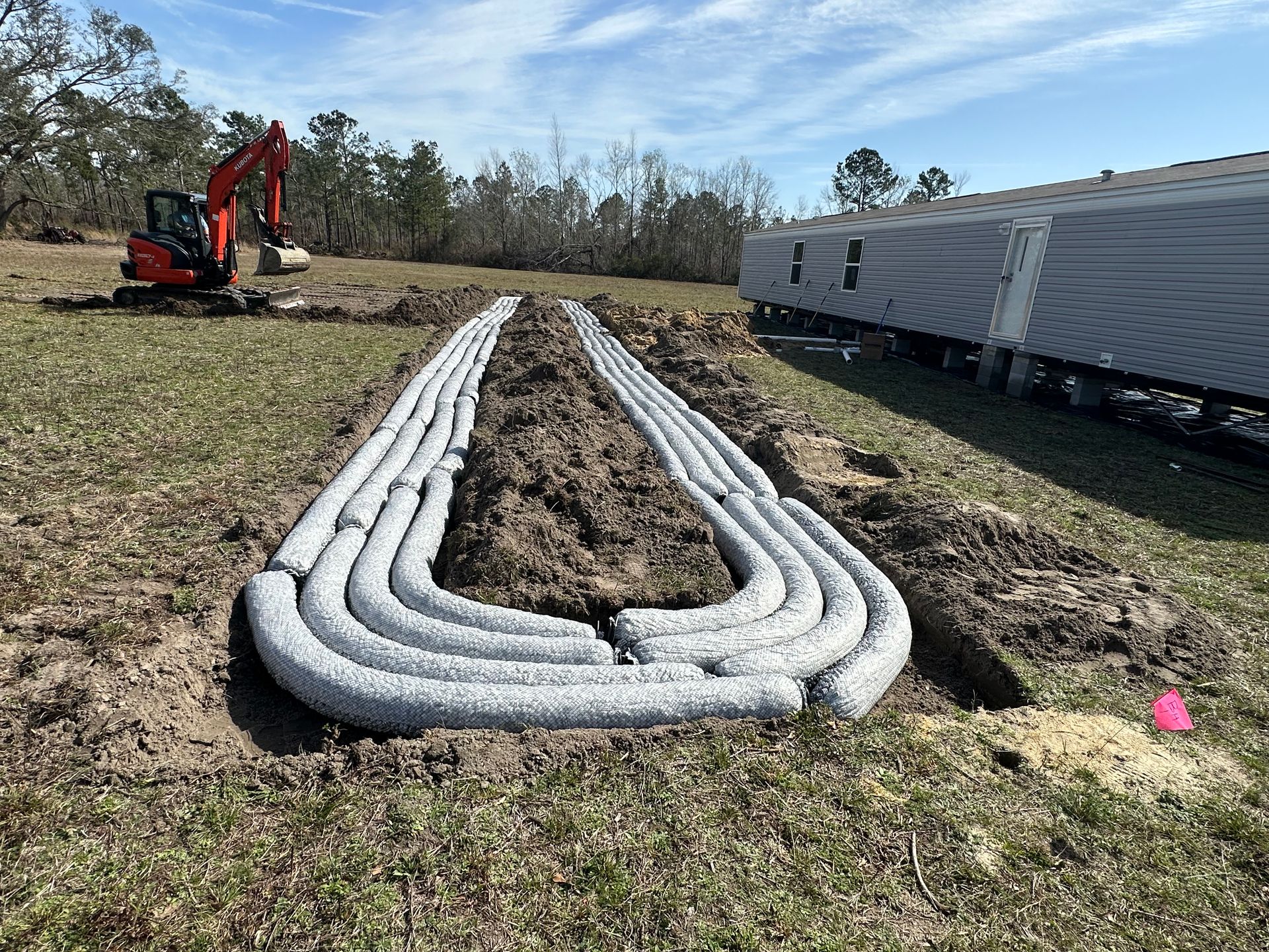 Excavator installing drainage pipes in a field beside a white trailer under a cloudy sky