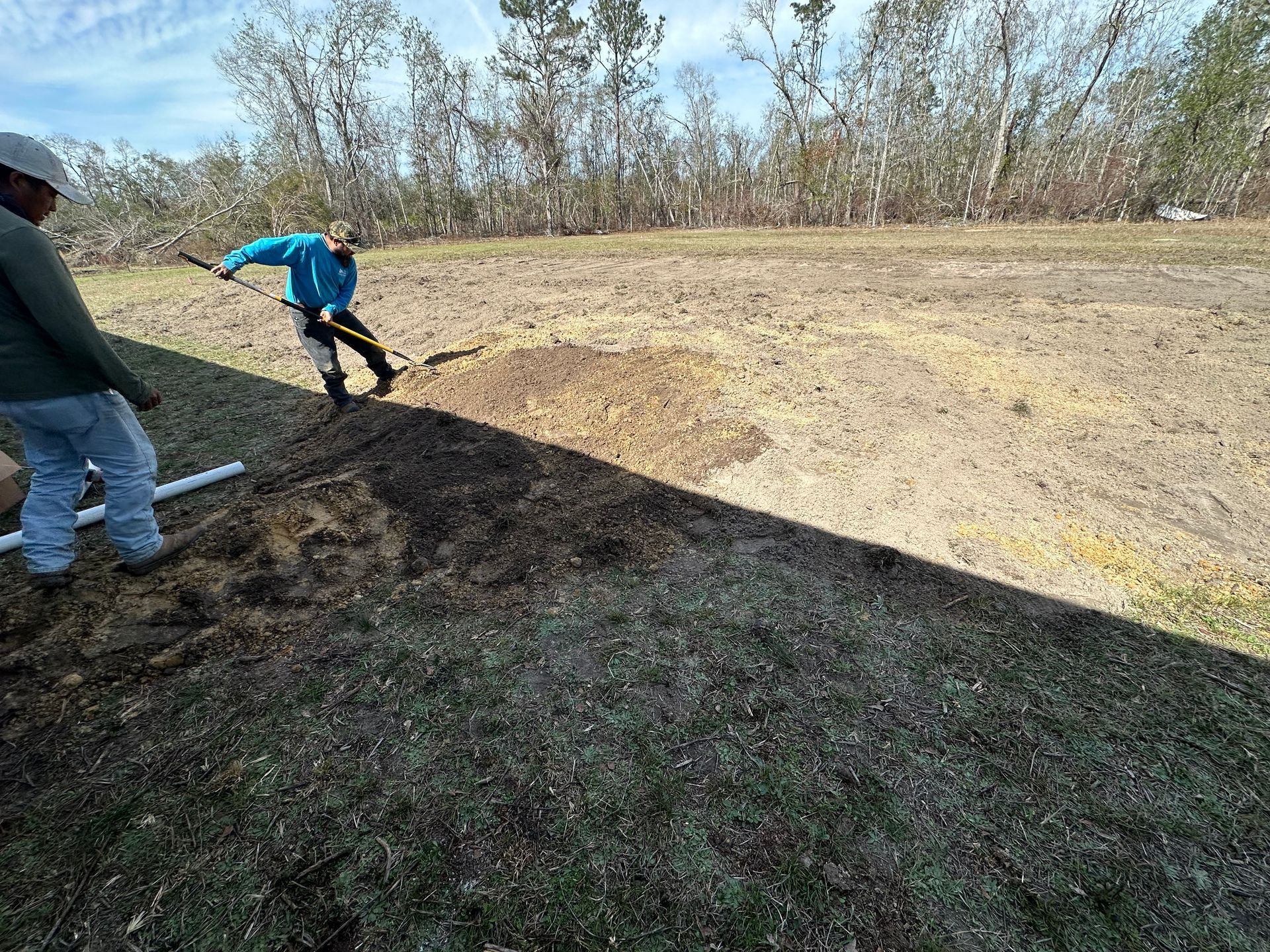 Two workers shaping a dirt berm beside a bare field on a sunny day