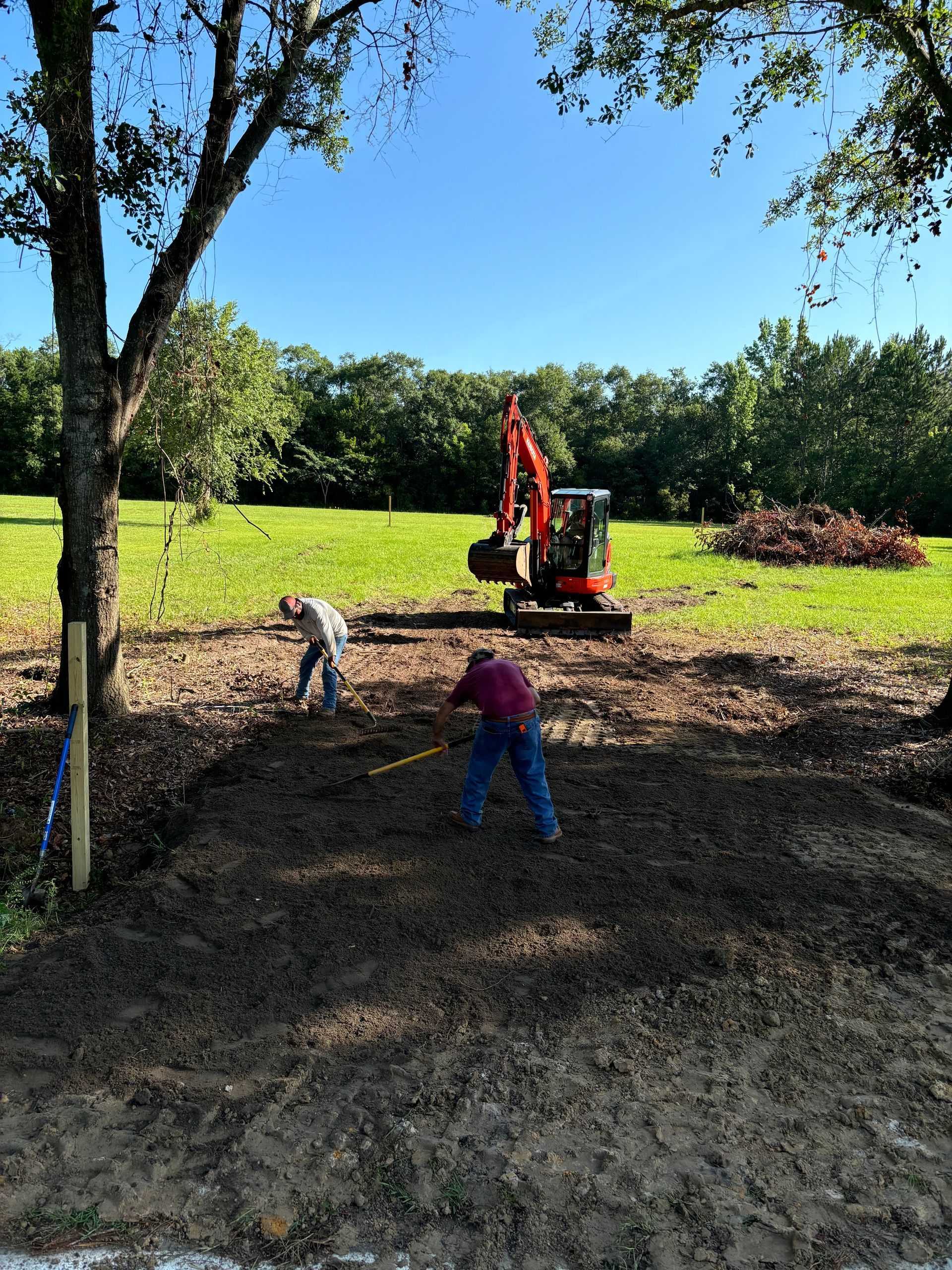 Two workers with an excavator clearing dirt in a sunny grassy field under trees