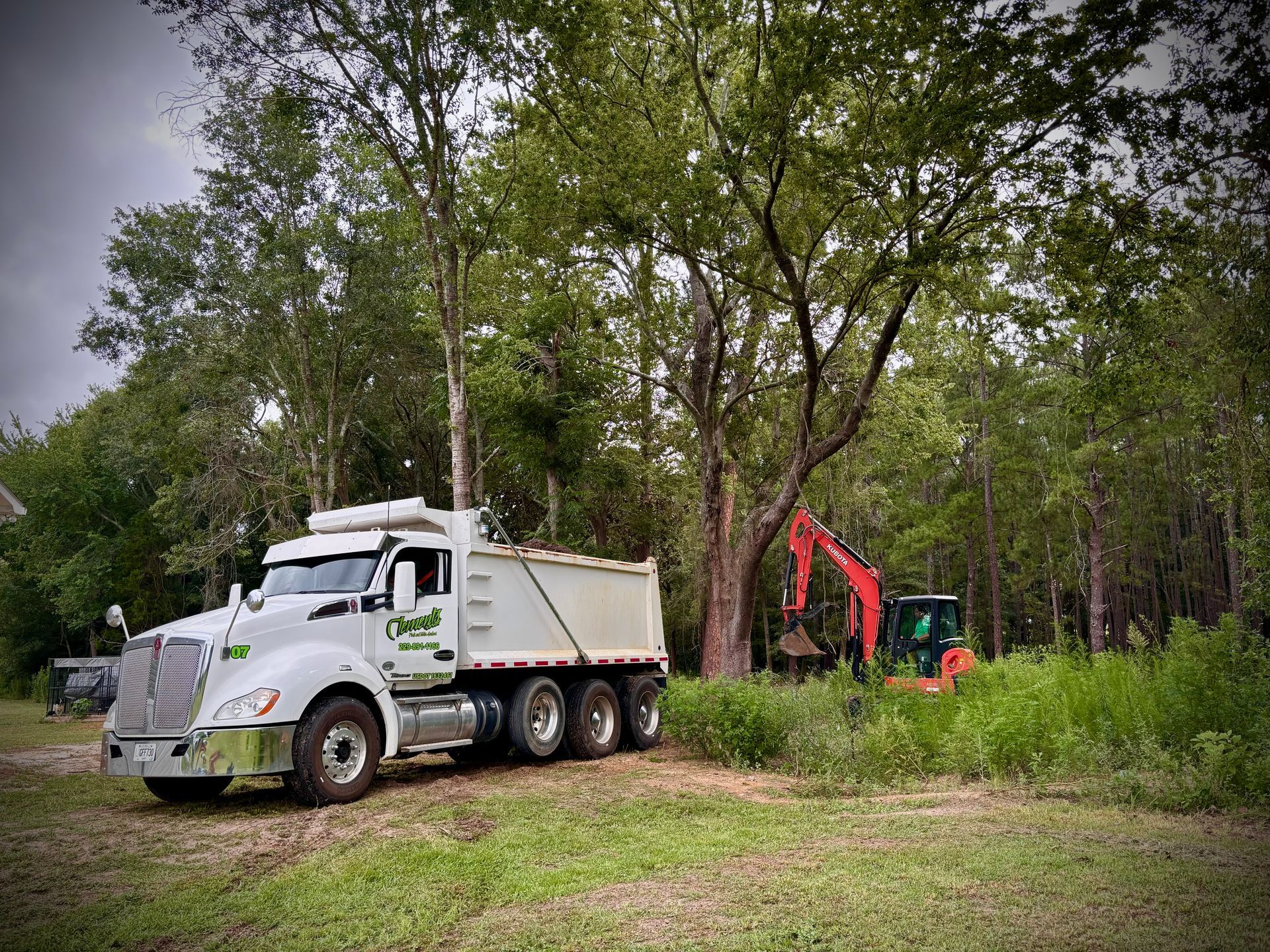 White dump truck and red excavator in a wooded clearing beside grass and trees