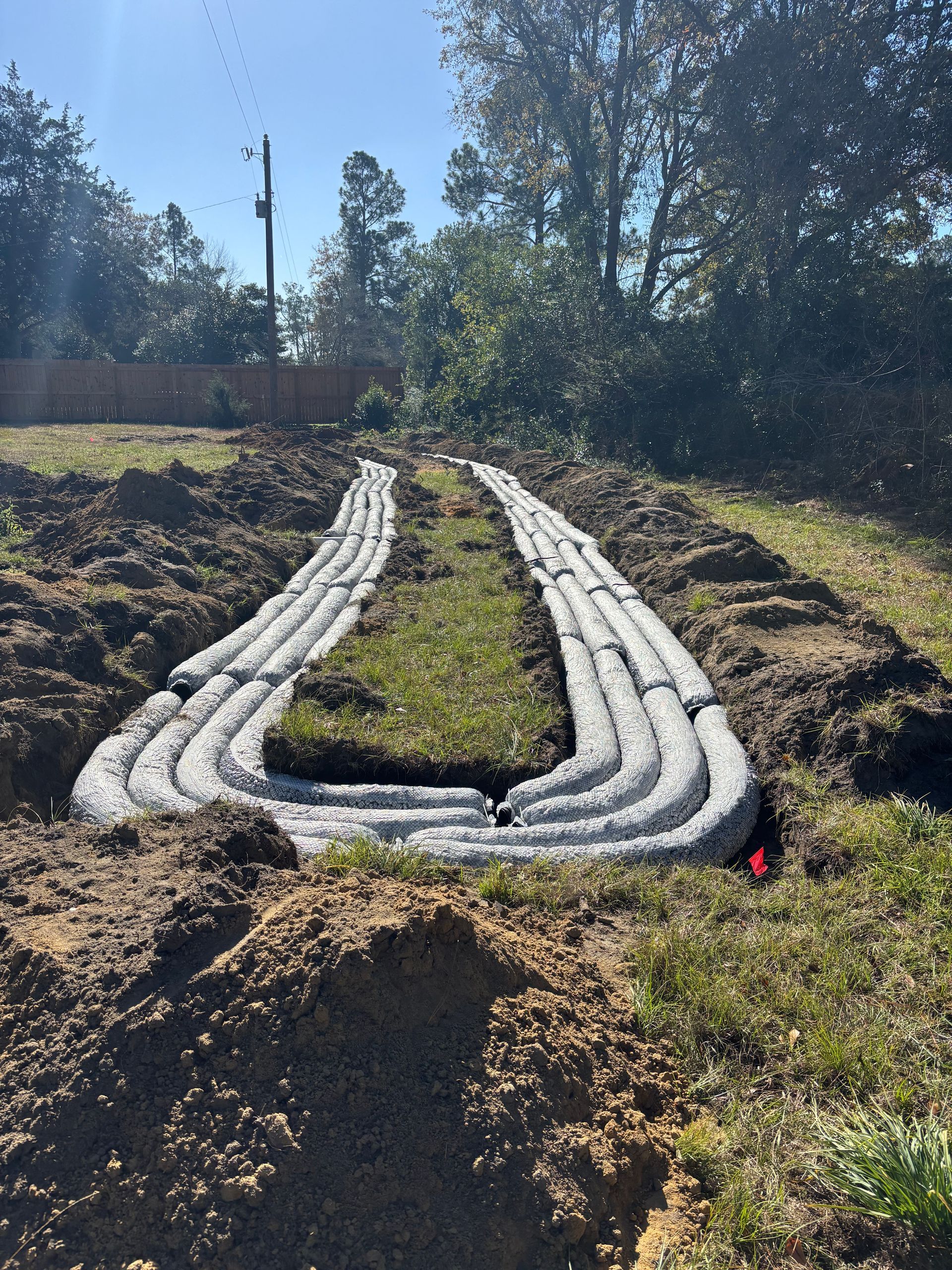 Long white pipe coils laid in a muddy trench on a grassy slope under trees.