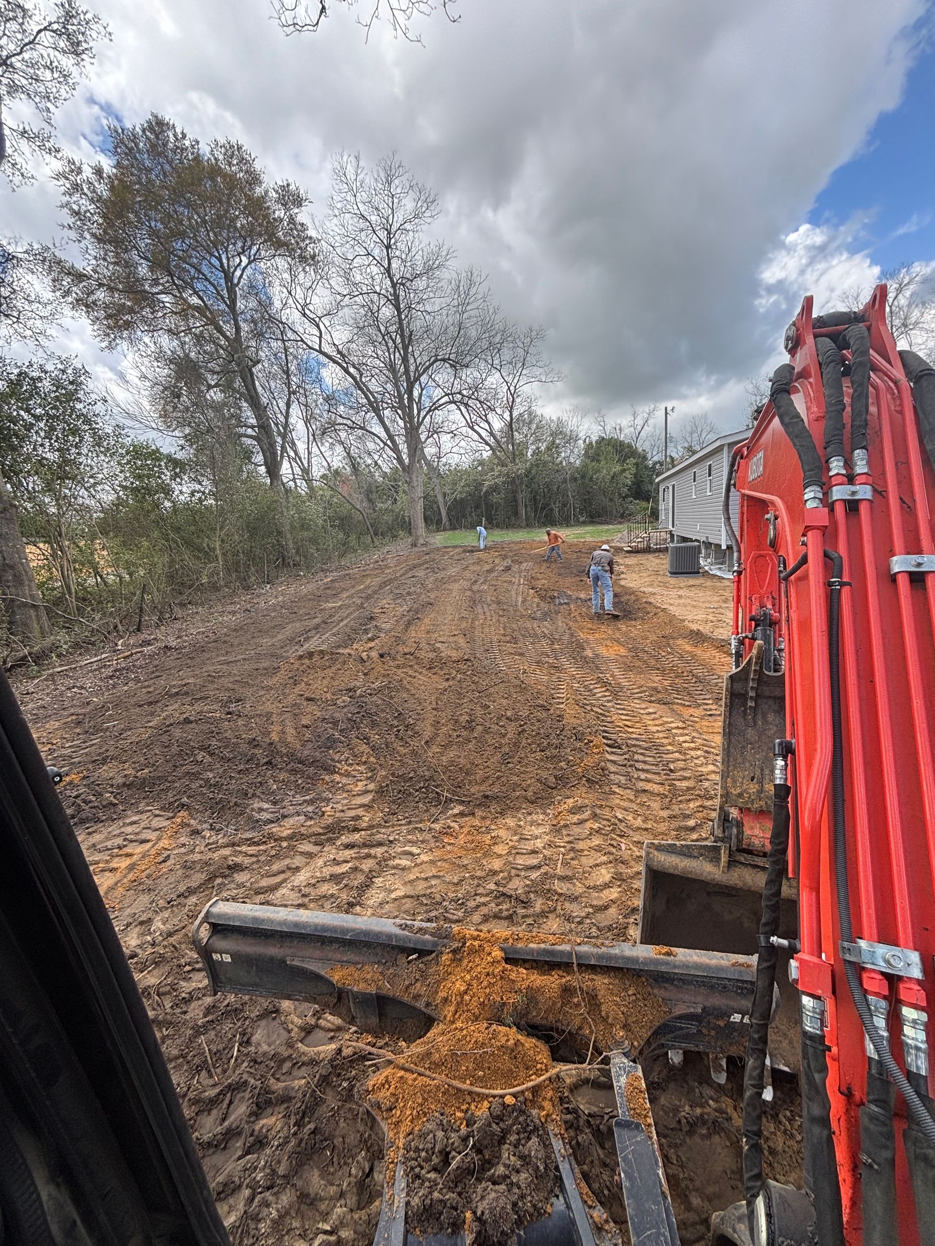 Construction excavator clearing a muddy hillside under cloudy skies, with red equipment at right.