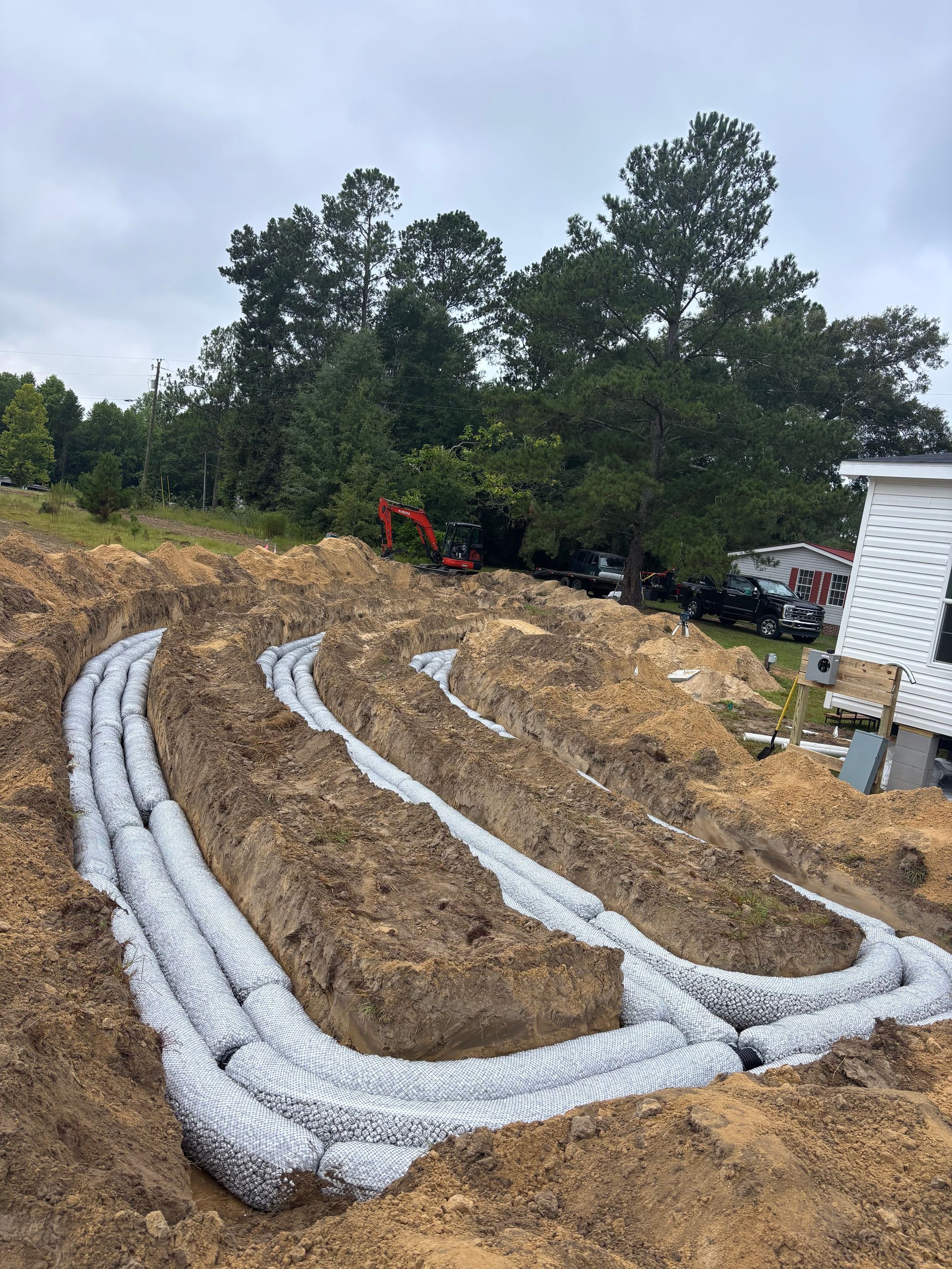White erosion-control tubes snake across a muddy hillside beside trees and a white building.