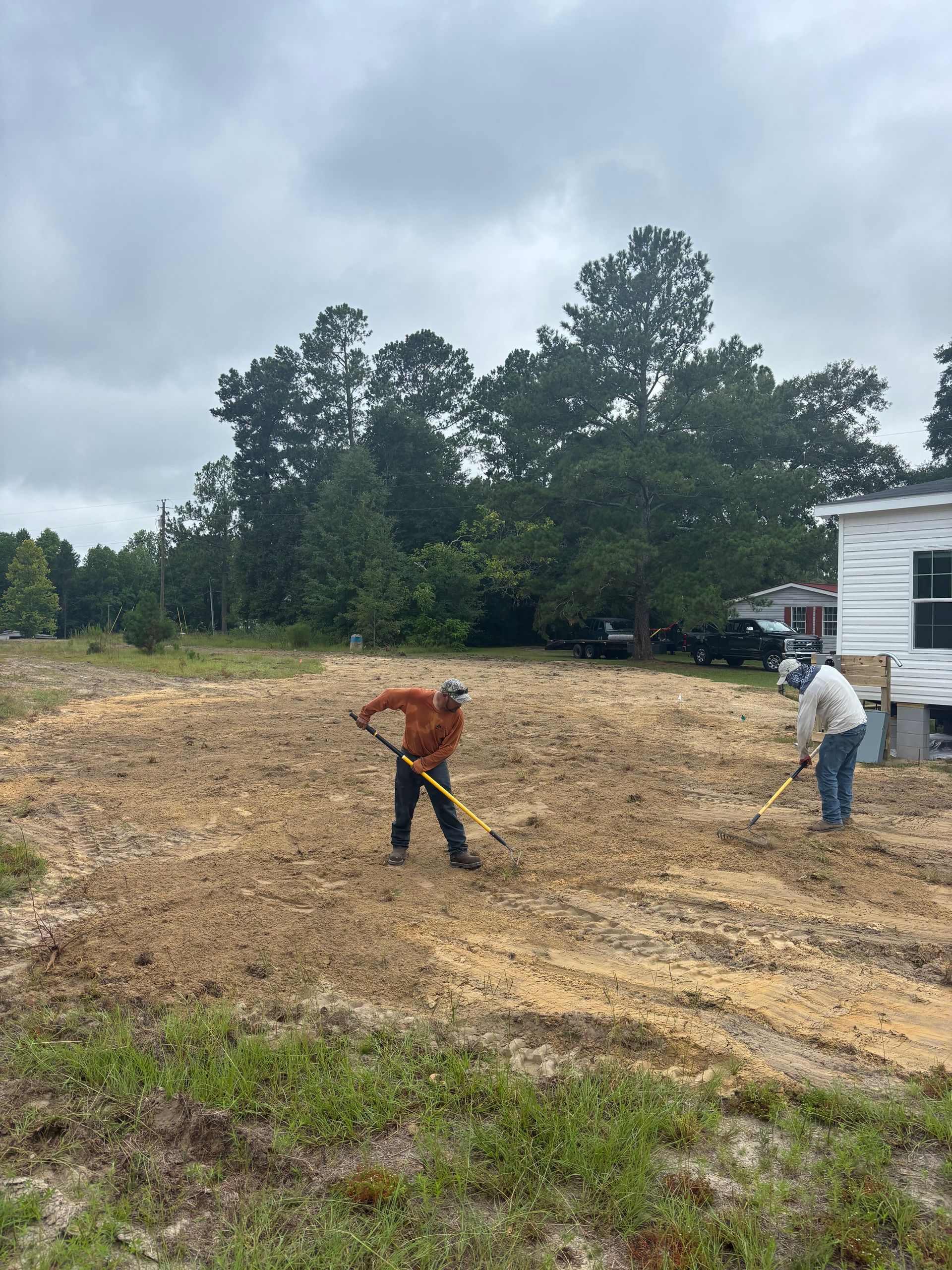 Two workers grading a dirt lot beside a white house under a cloudy sky