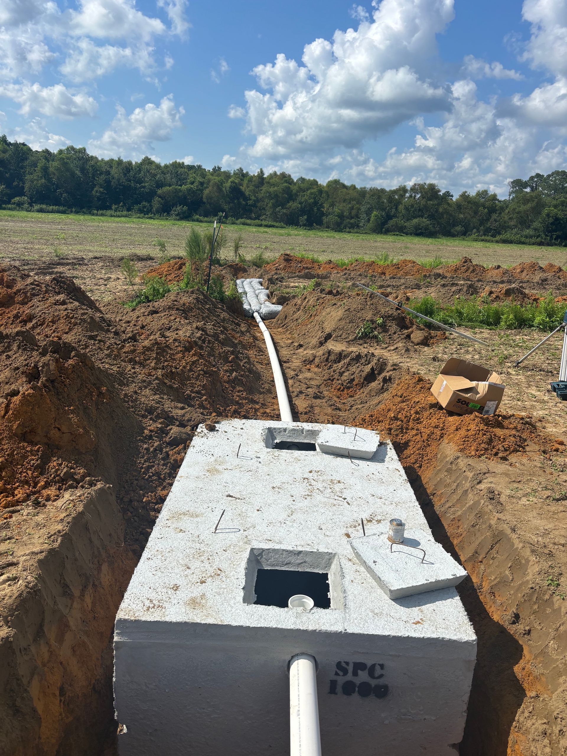 Concrete utility chamber in a dirt trench with a white pipe, in a field under a cloudy sky