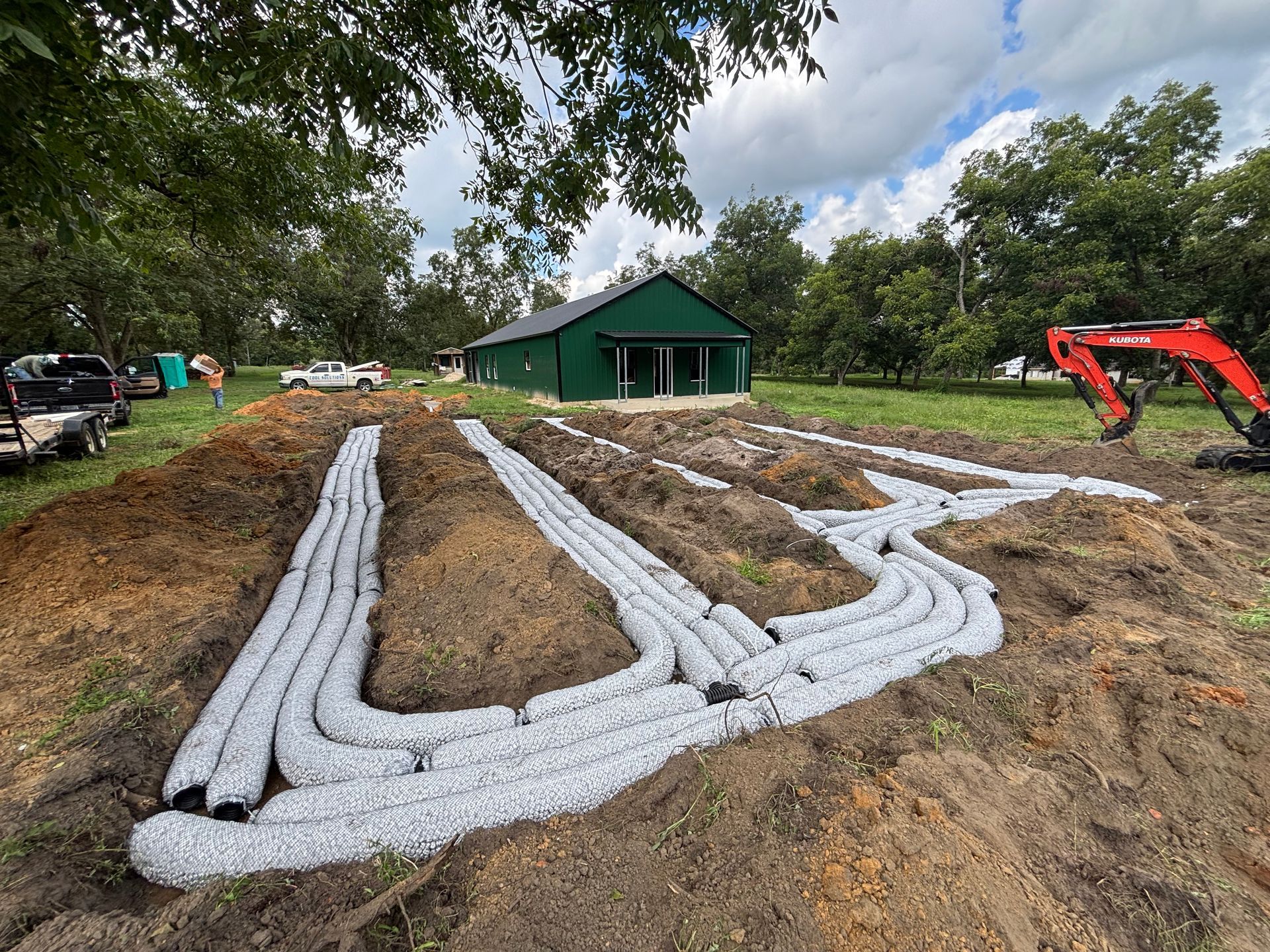 Crawlspace drainage installation with white pipe trenches beside a green house and excavator in a dirt yard