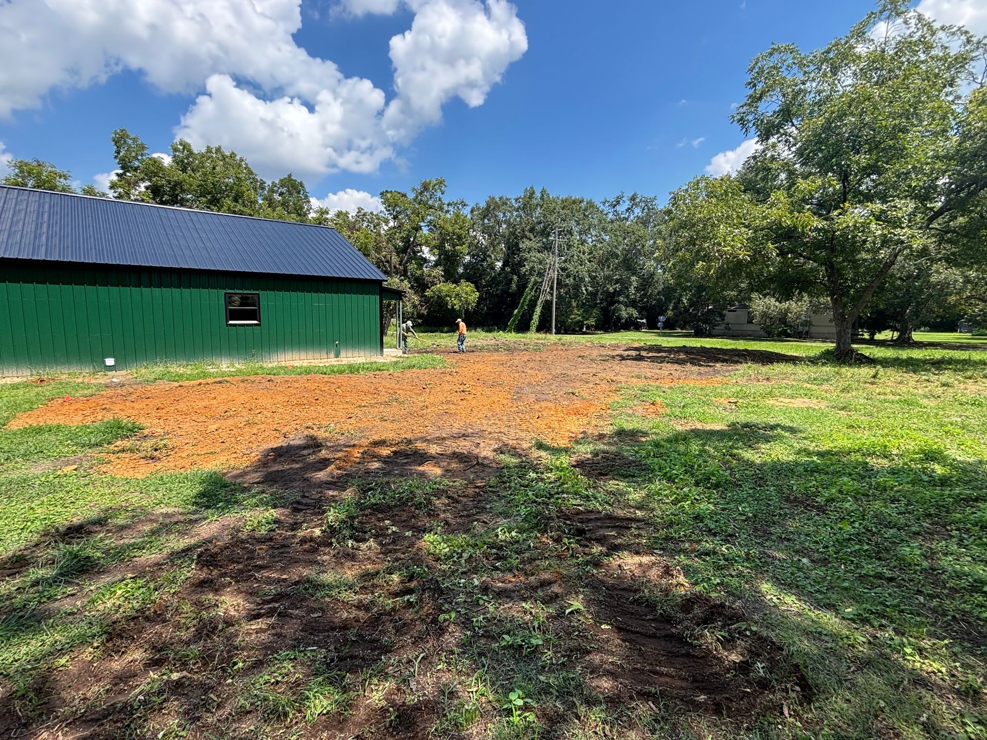 Green barn beside a cleared orange-brown field under a blue sky with clouds, trees in the background