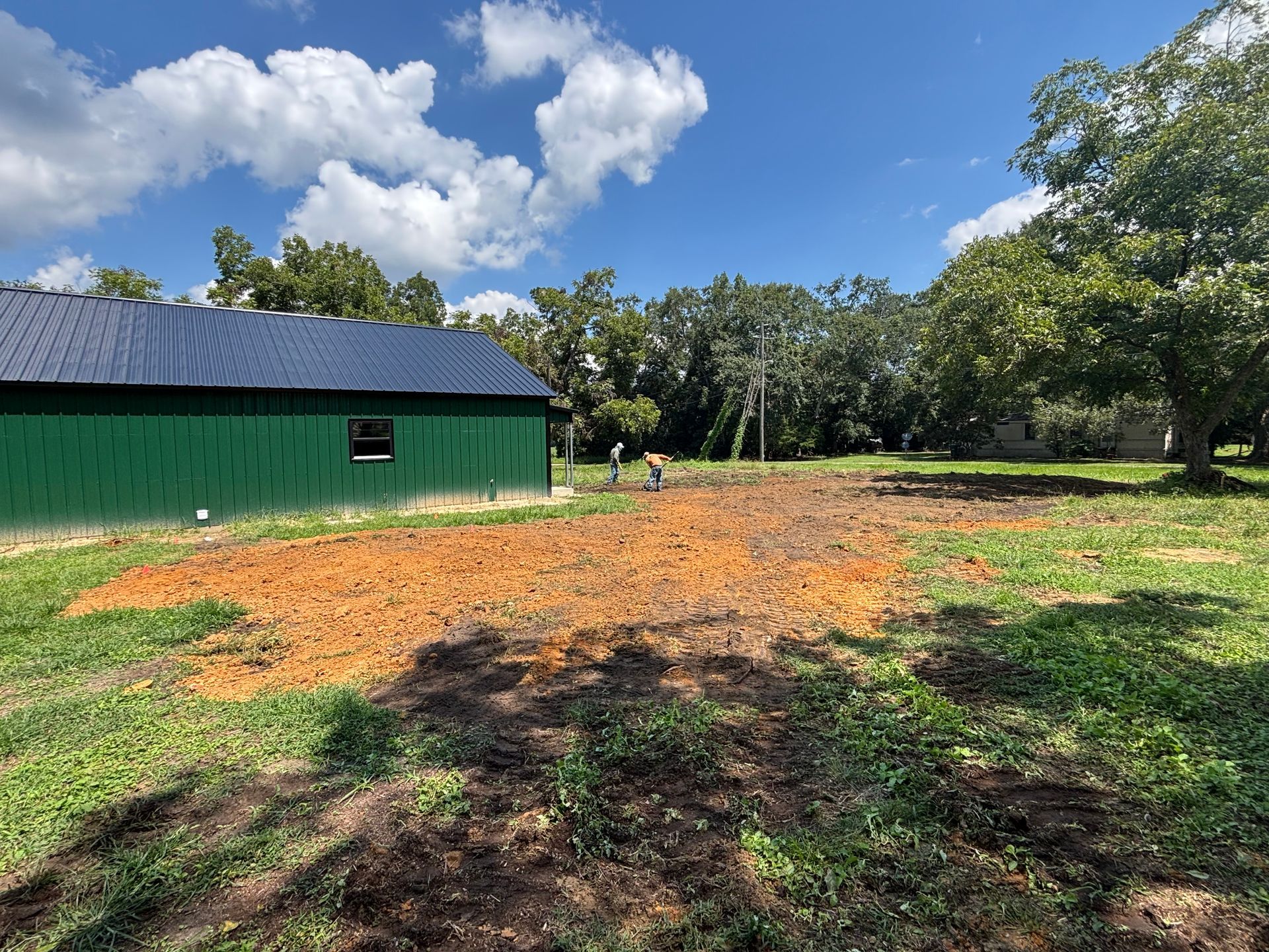 Green barn beside a freshly cleared orange dirt patch under a bright blue sky with clouds