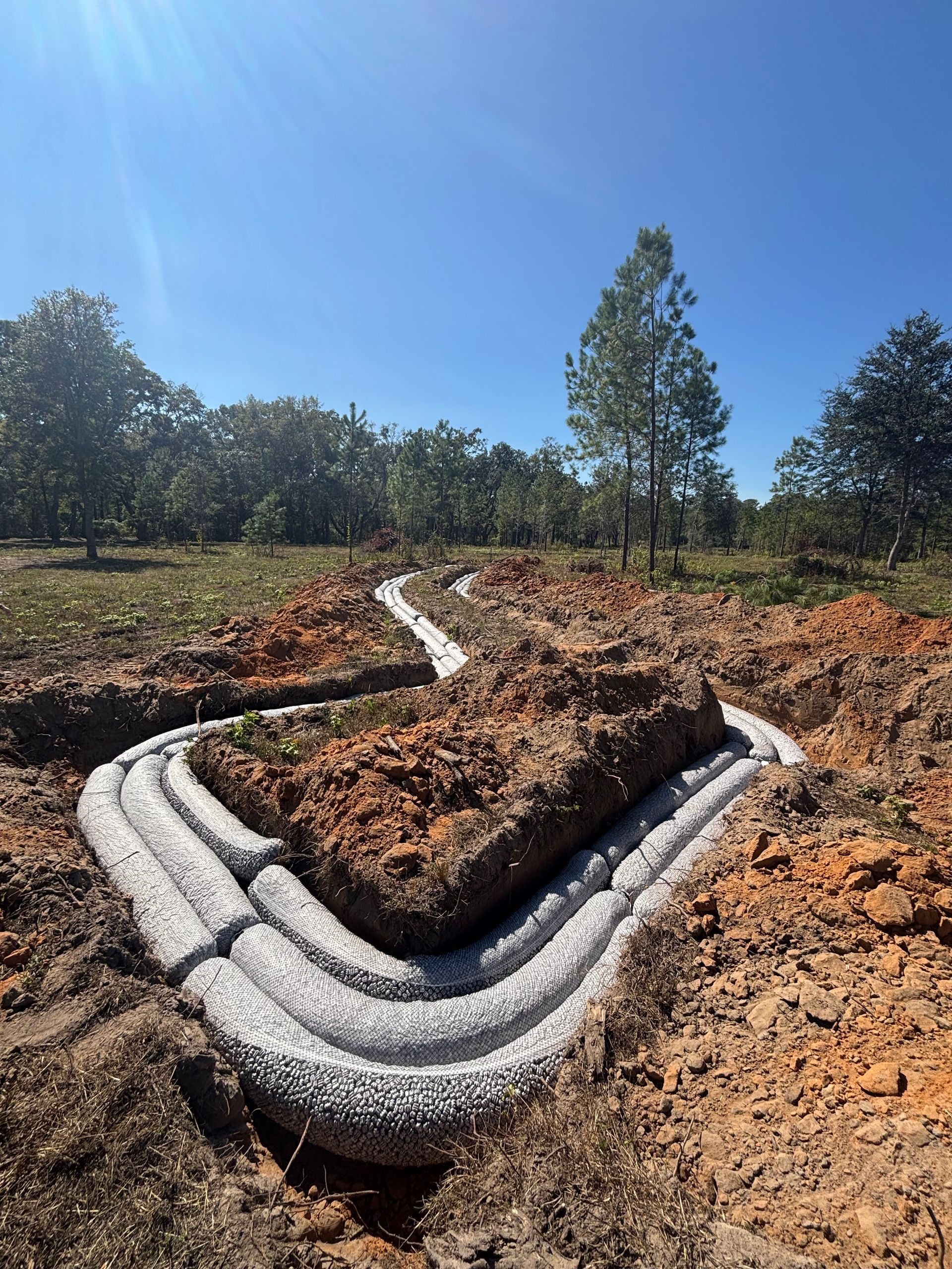 White plastic erosion-control pipes forming a winding trench in a dry, grassy field under a blue sky