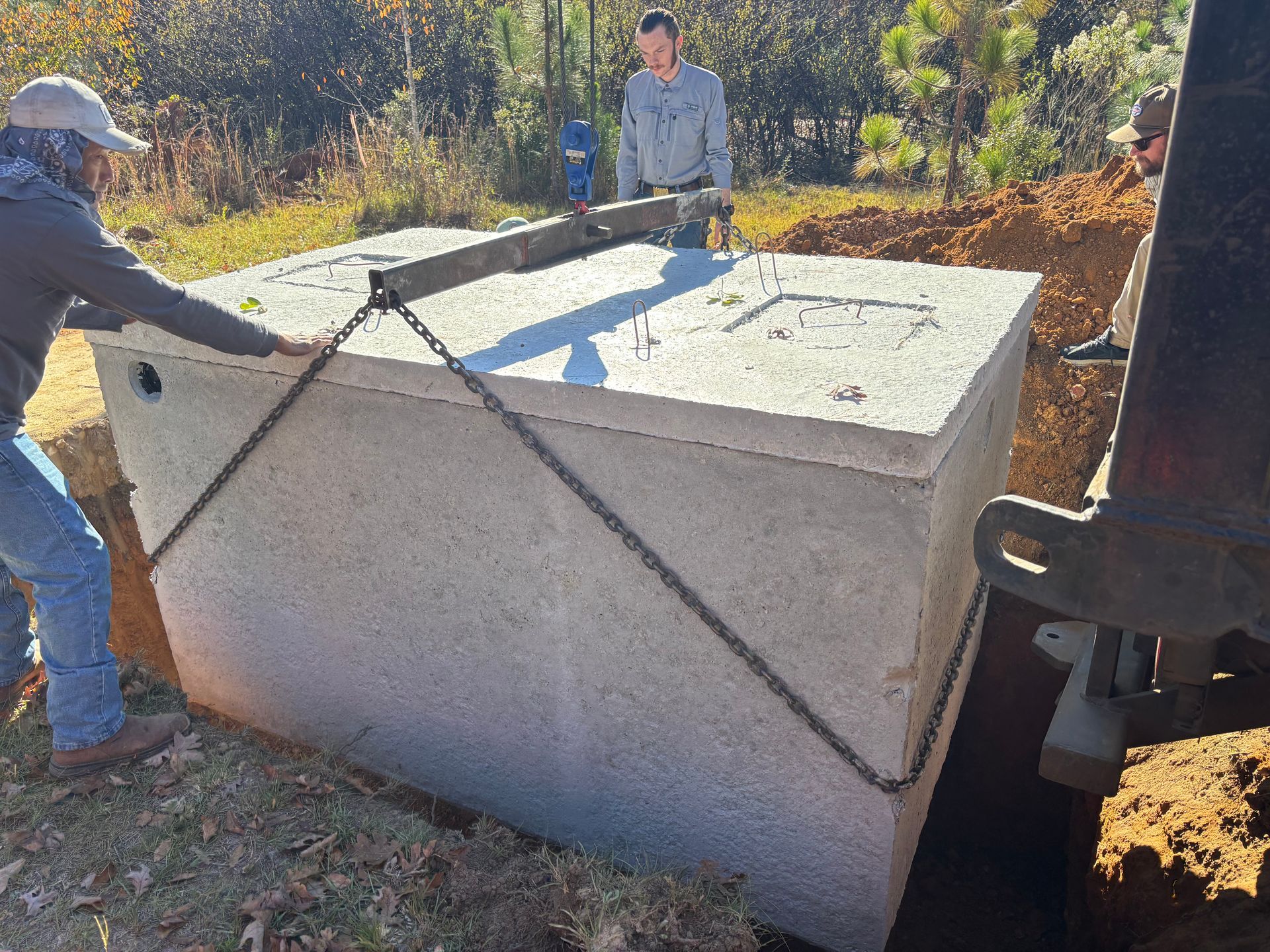 Workers lowering a concrete utility box into a dug hole with straps outdoors