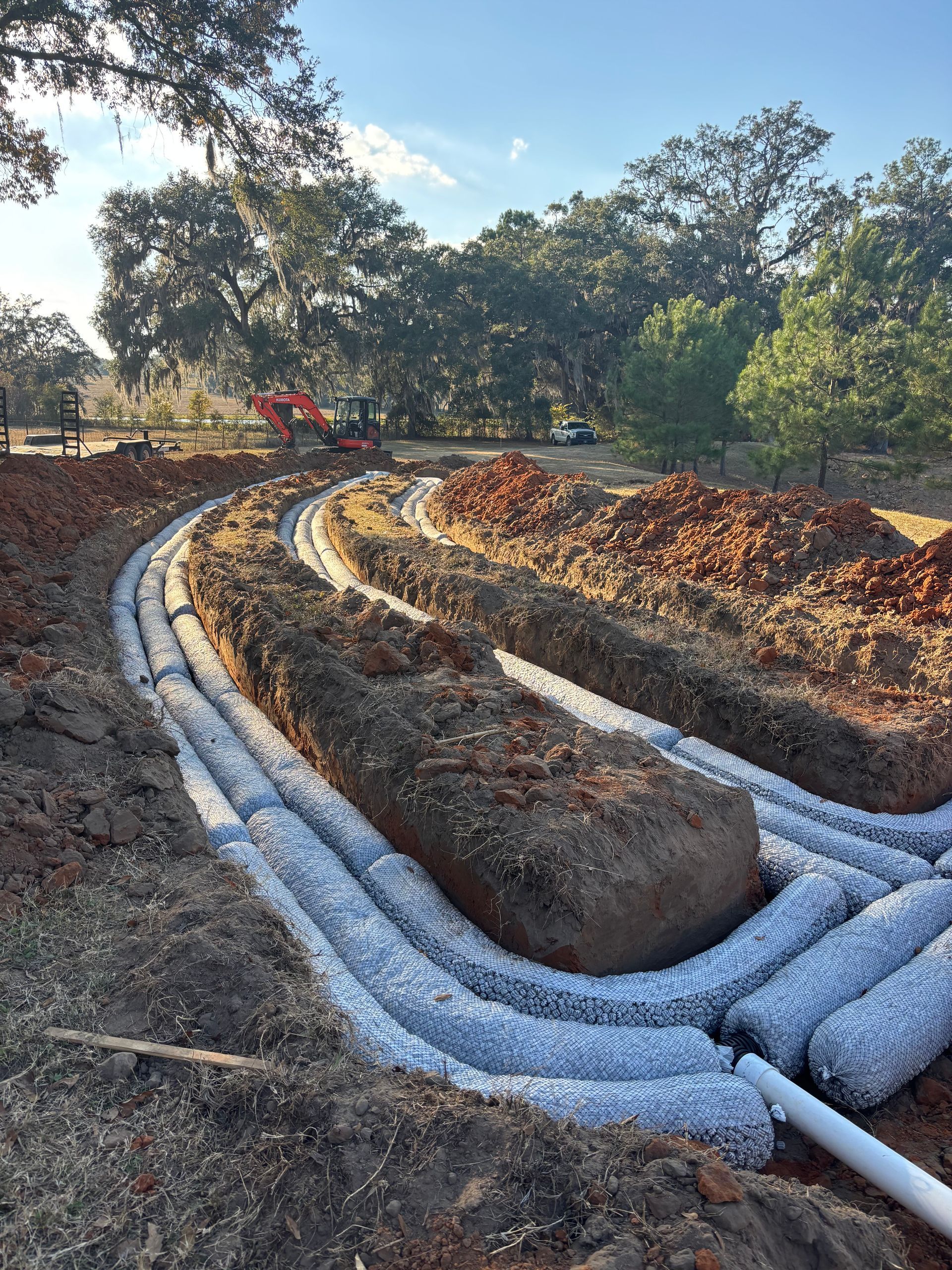 Curved drainage pipes lined with white gravel in a sunlit dirt excavation site with trees in the background