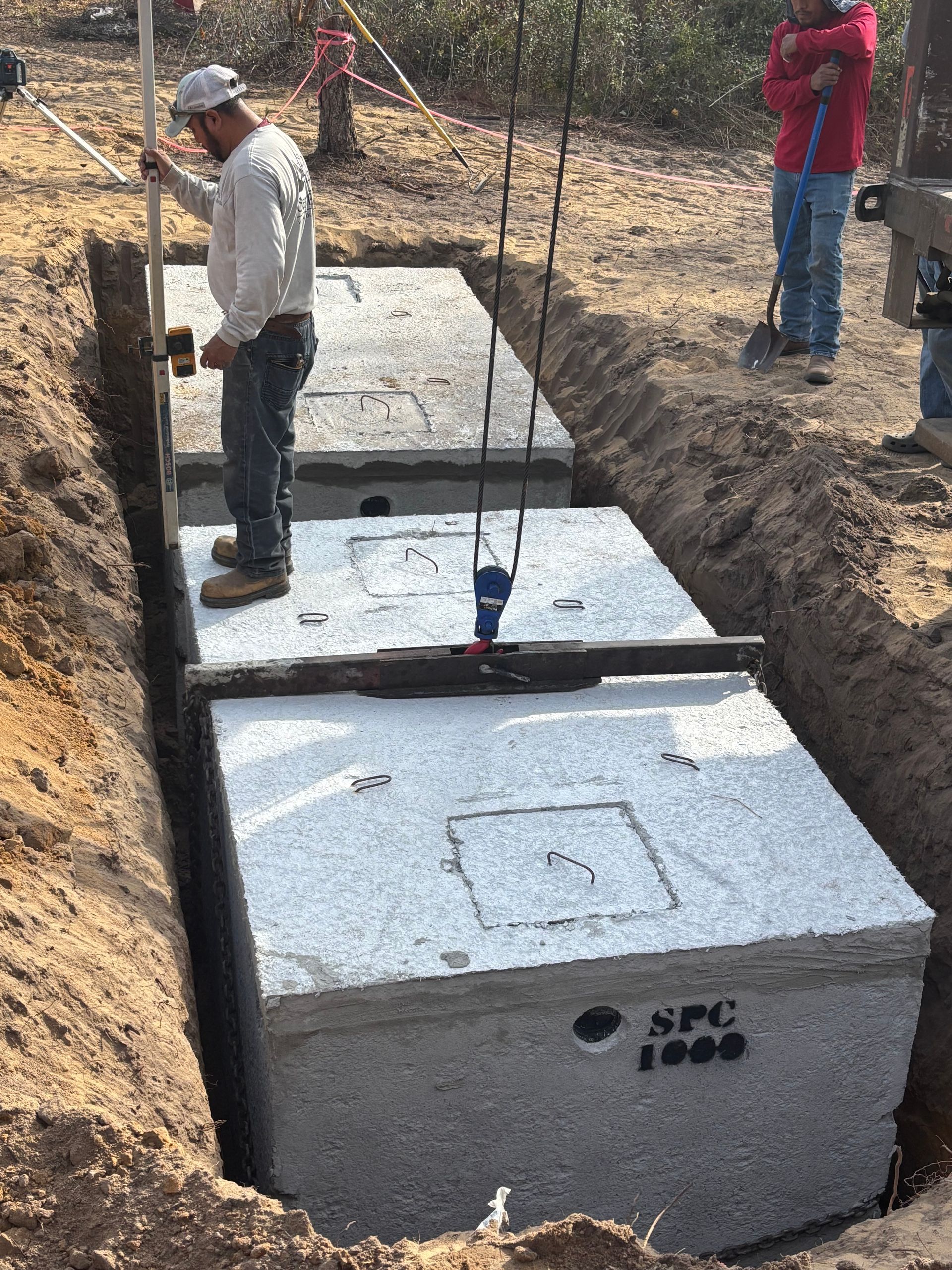 Workers installing large concrete septic tanks in a trench at a construction site.