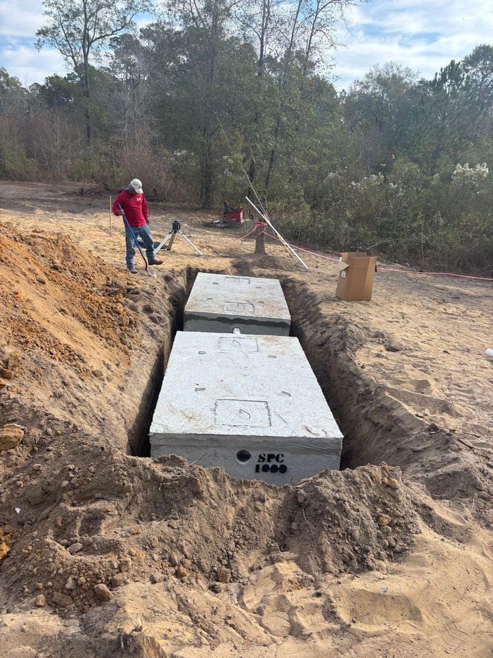 Two rectangular concrete septic tanks in a trench with a worker nearby, surrounded by dirt and trees.