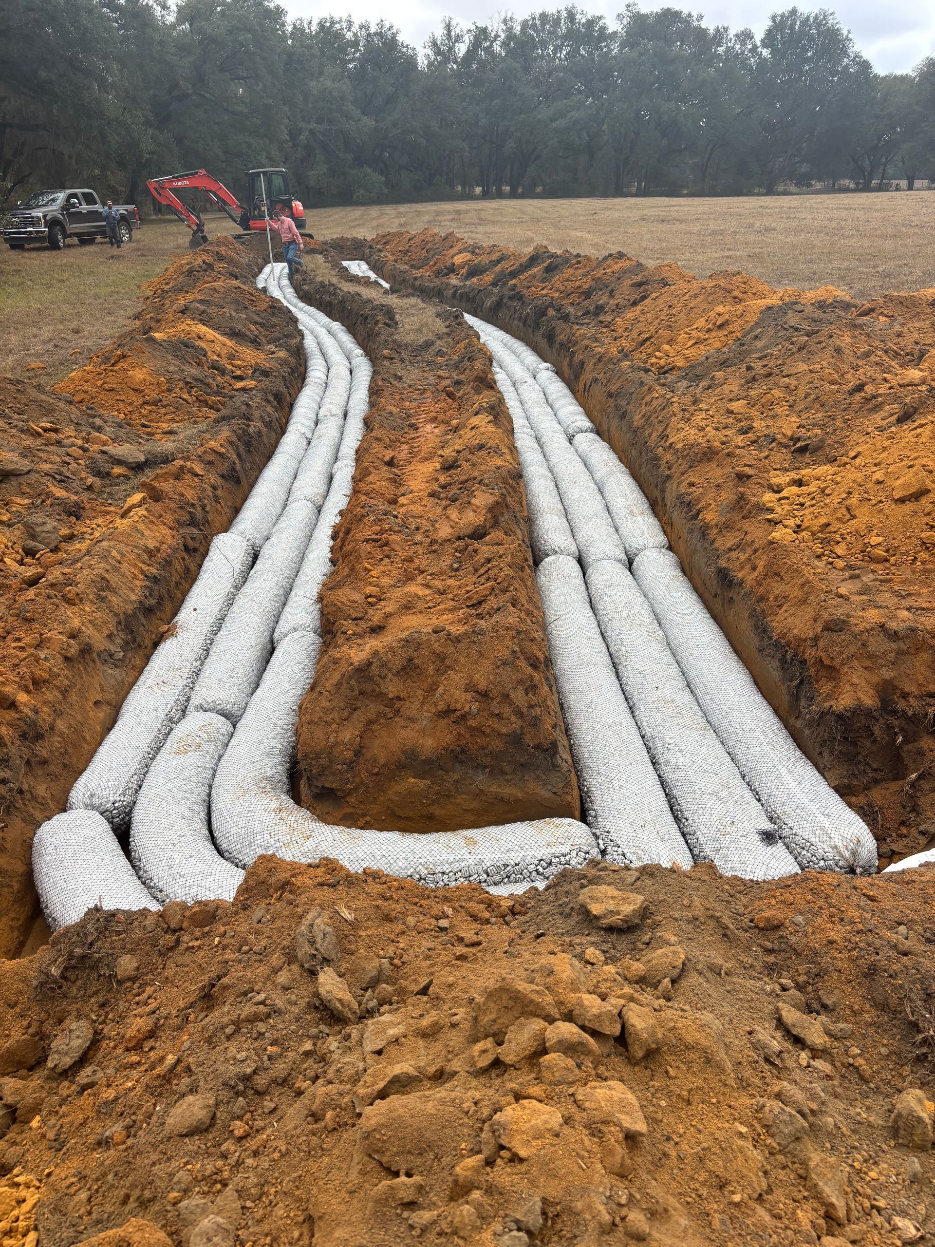 Mud-covered white drainage pipes laid in trenches at a construction site under overcast skies