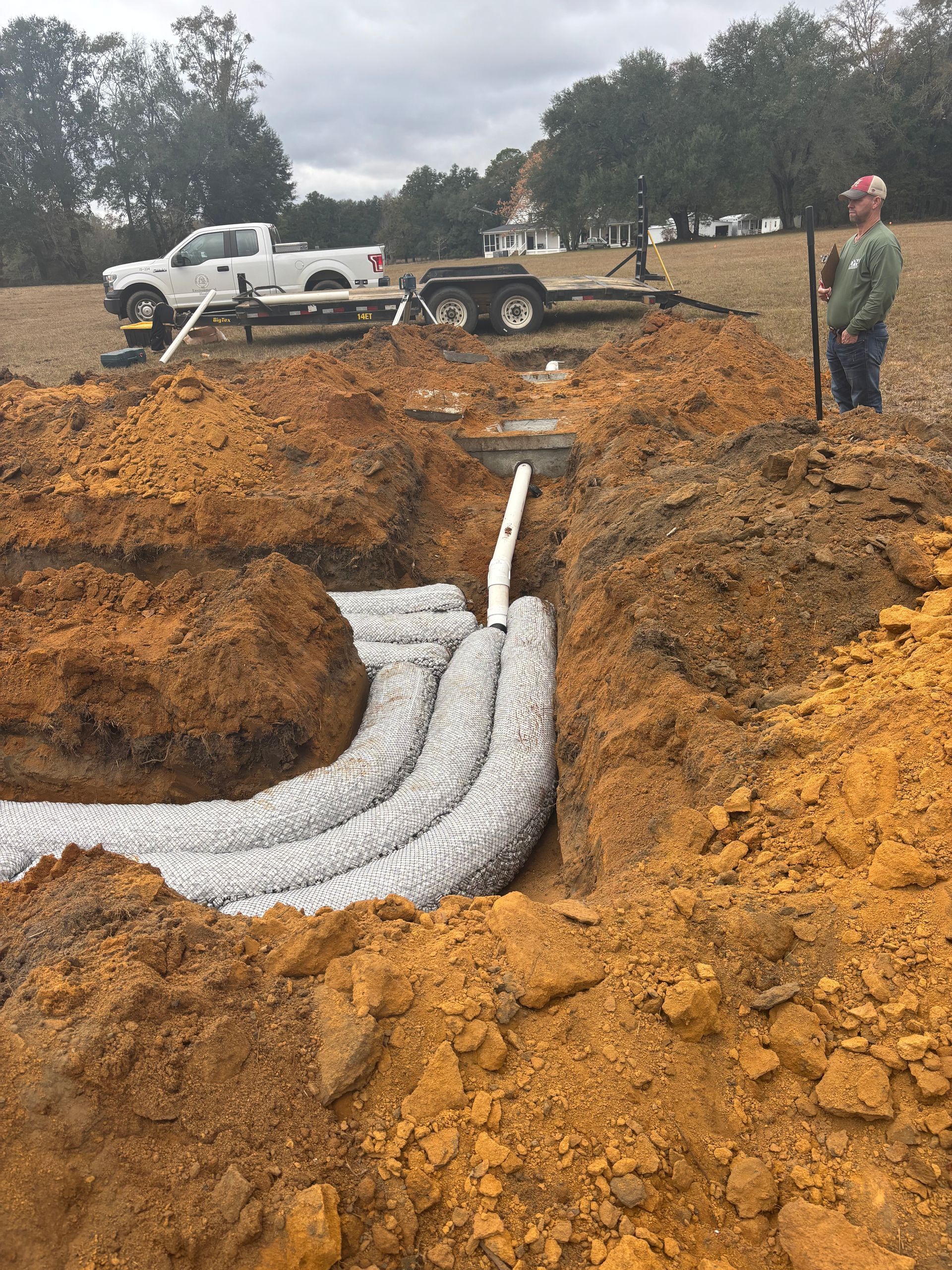 Workers installing white drainage pipe in a muddy trench at a construction site.