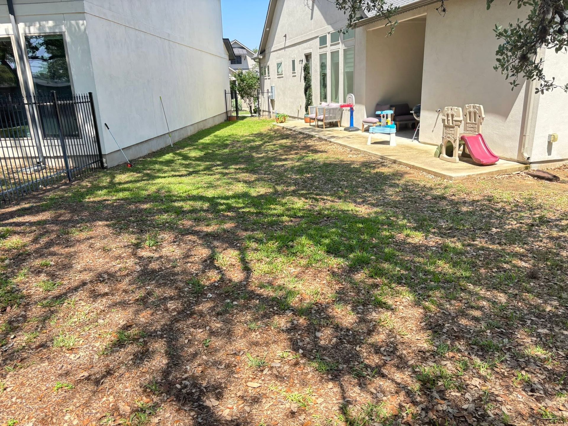 Narrow backyard with patchy grass, trees casting shadows, and a house wall lined with children’s toys