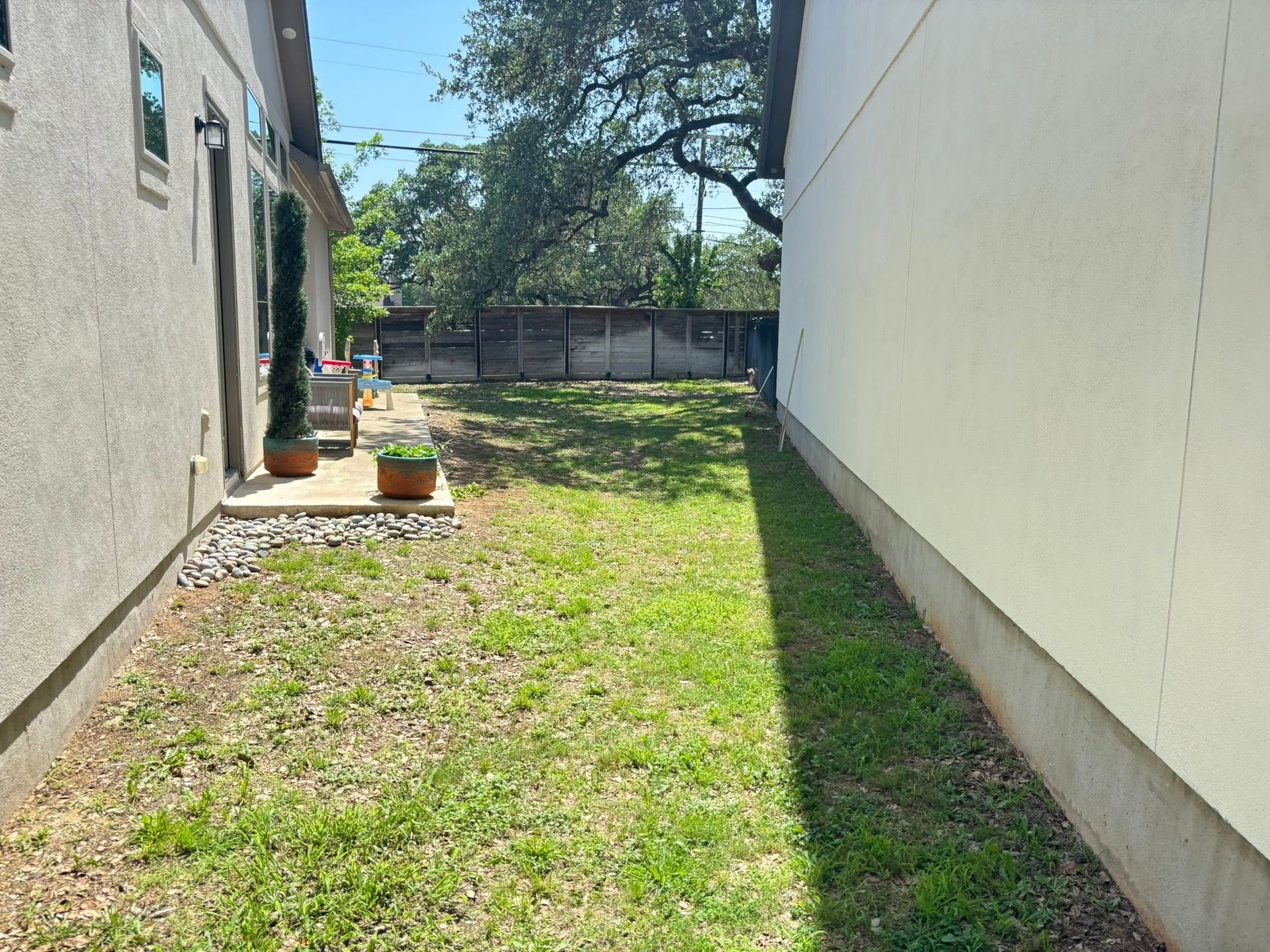 Narrow grassy side yard between two buildings, with potted plants and a fence at the back.