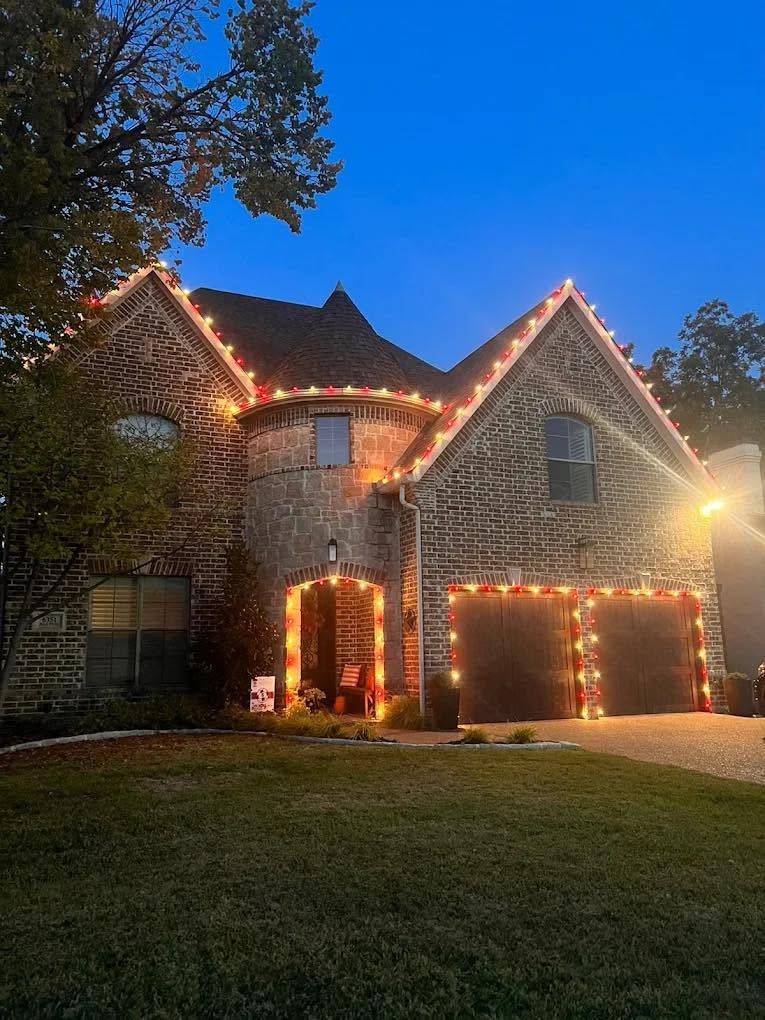 Two-story house at dusk outlined with warm holiday lights, with illuminated garage and front doorway