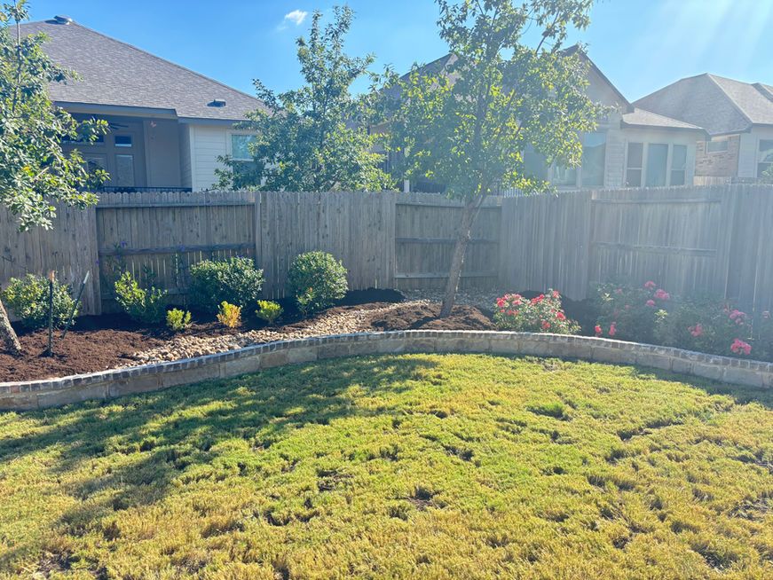 Sunny backyard with green lawn, stone flower bed, wooden fence, and trees behind a house