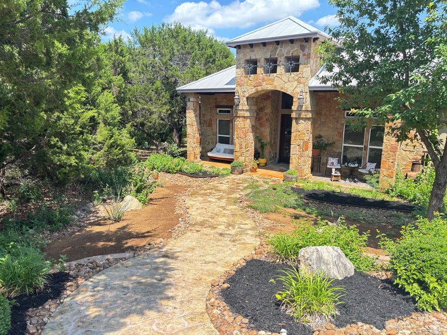 Stone house entrance with arched doorway, metal roof, and landscaped garden path surrounded by trees
