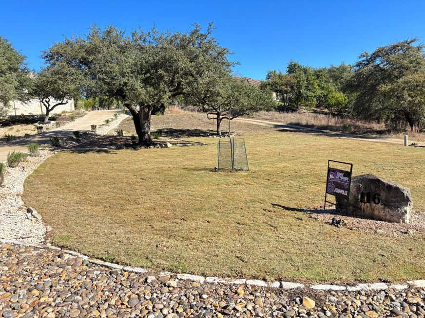 Dry landscaped park with trees, gravel paths, and a stone memorial marker under a clear blue sky