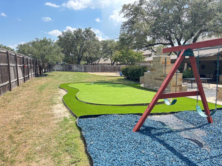Outdoor playground with green turf, blue mulch, a red swing frame, and a fenced yard under a sunny sky