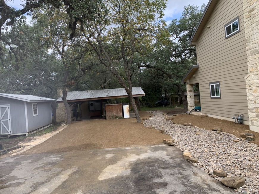Driveway and carport beside a beige house, with gravel landscaping and trees overhead.