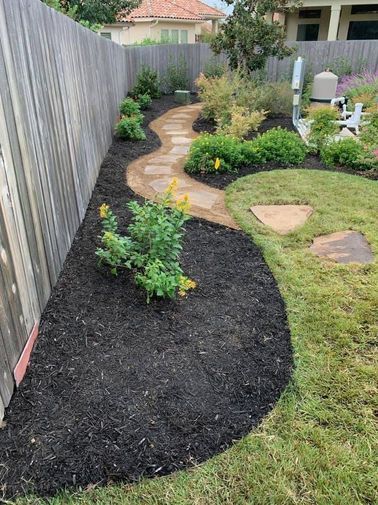Curved garden bed with fresh mulch, small shrubs, stepping stones, and lawn beside a wooden fence.