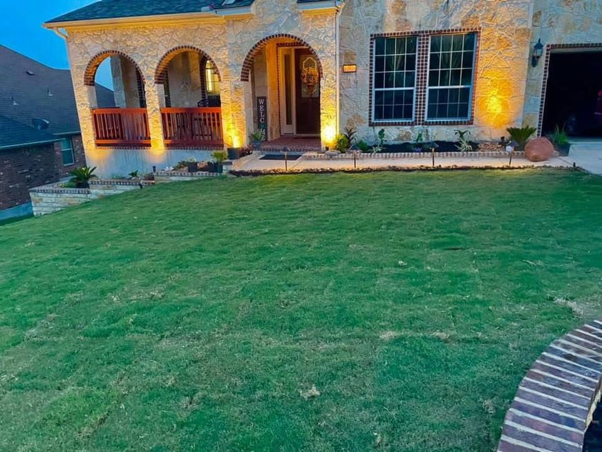 Stone house with arched porch, lit windows, and a large green lawn at dusk.