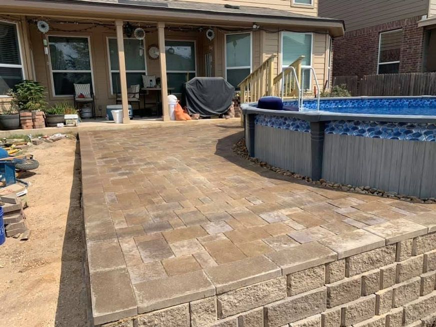 Backyard patio with tan pavers leading to a raised above-ground pool beside a house.