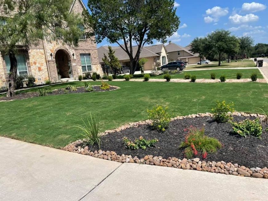 Suburban front yard with green lawn, flower beds, stone house, and concrete driveway under a sunny sky
