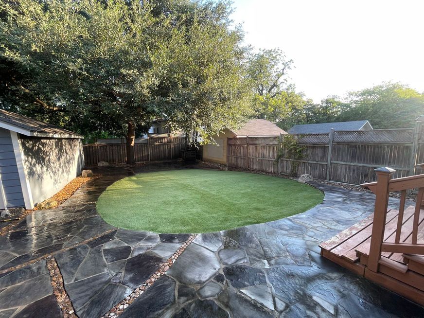 Backyard with stone patio, circular artificial grass area, wooden deck, and fenced trees in the background
