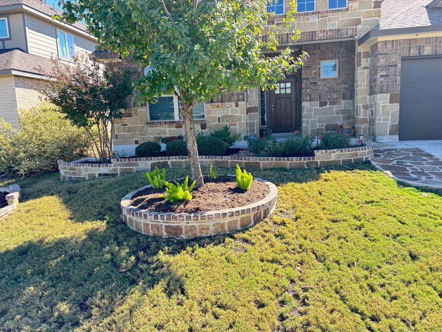 Front yard with stone house, circular brick planter, and green lawn in sunlight