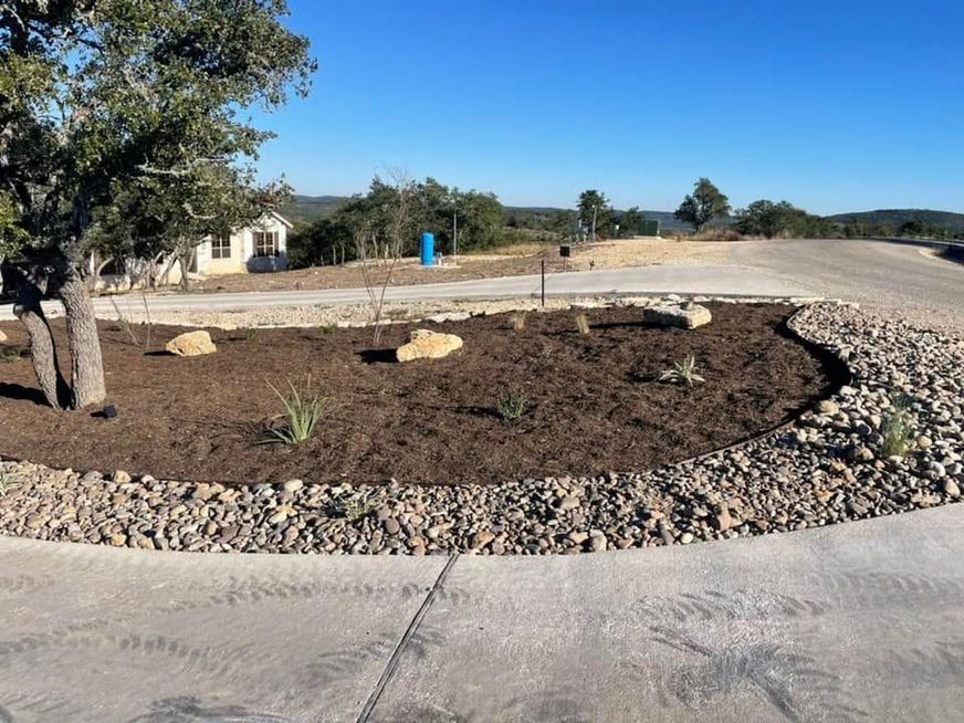 Landscaped roadside median with dark mulch, rocks, and a young tree under a blue sky