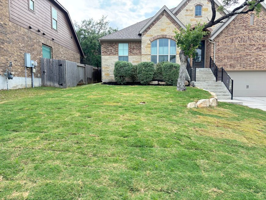 Suburban front yard with green lawn, brick house, bushes, and a driveway on the right.