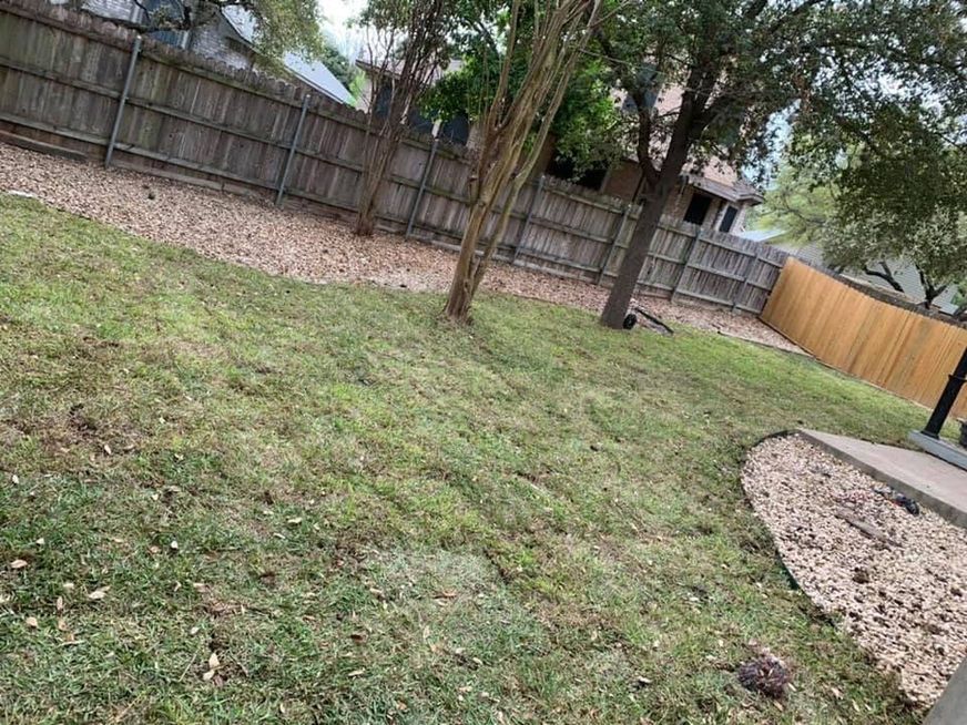 Backyard with grass, a tree, wooden fence, and a gravel border beside a patio.