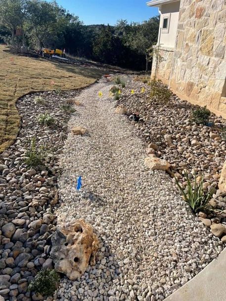Gravel garden path beside a stone house, bordered by rocks and small plants in sunlight