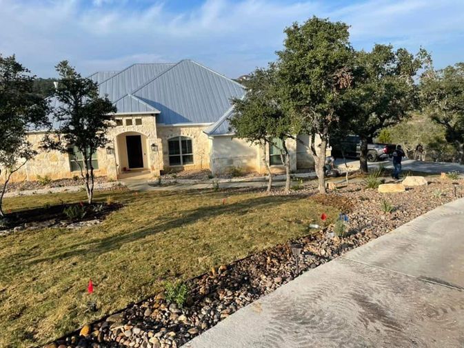 House with gray metal roof on a sunny lot, bordered by trees, grass, and a gravel road.