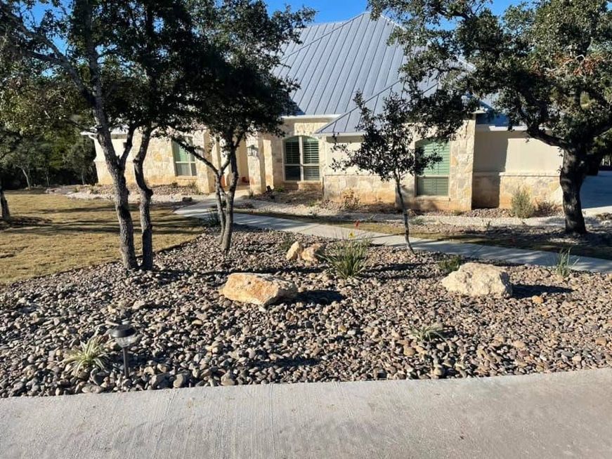 Single-story house with a rock garden, trees, and a gravel yard in bright daylight