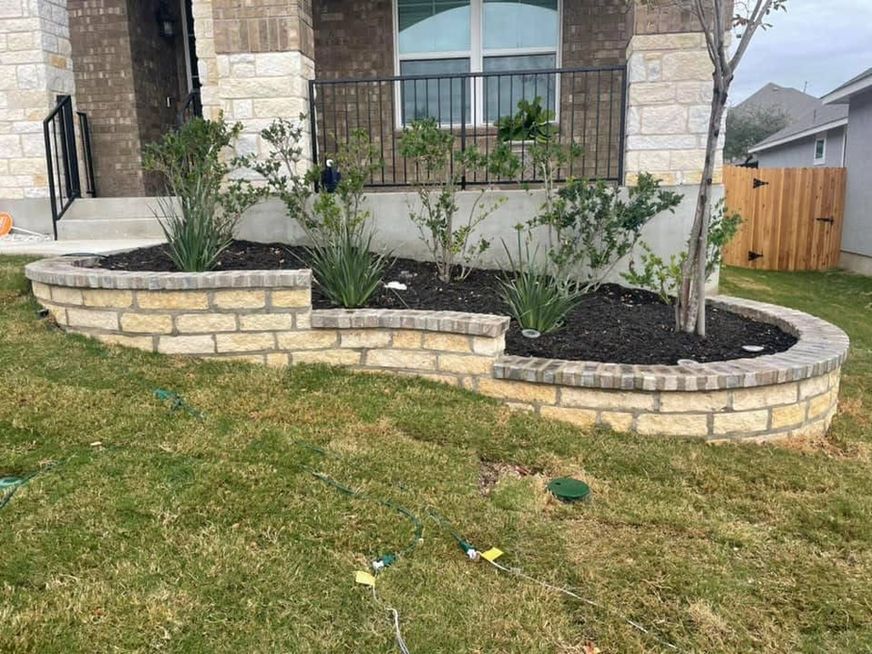 Stone house front yard with curved stacked-stone flower beds and young shrubs along the porch