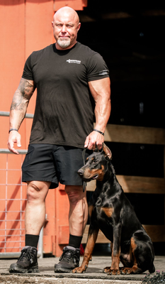 A muscular man in a black t-shirt and shorts standing beside a sitting black and tan Doberman dog against a red wall.